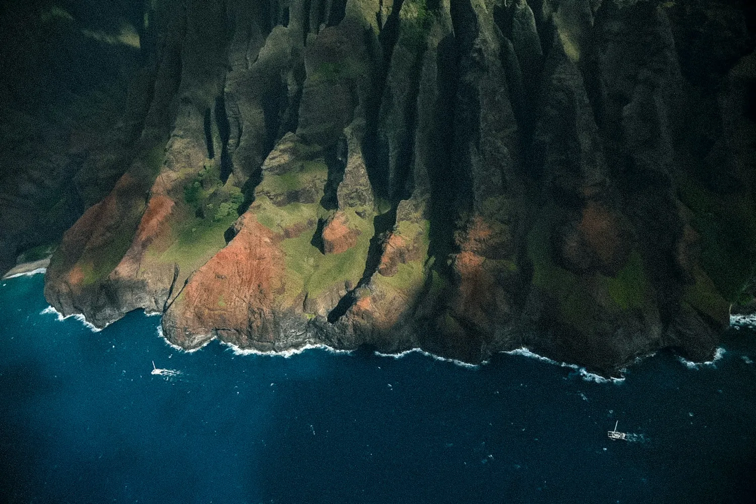 Looking down on deep-green fluted Nāpali cliffs meeting dark blue water, with two small boats near the foot of the rock