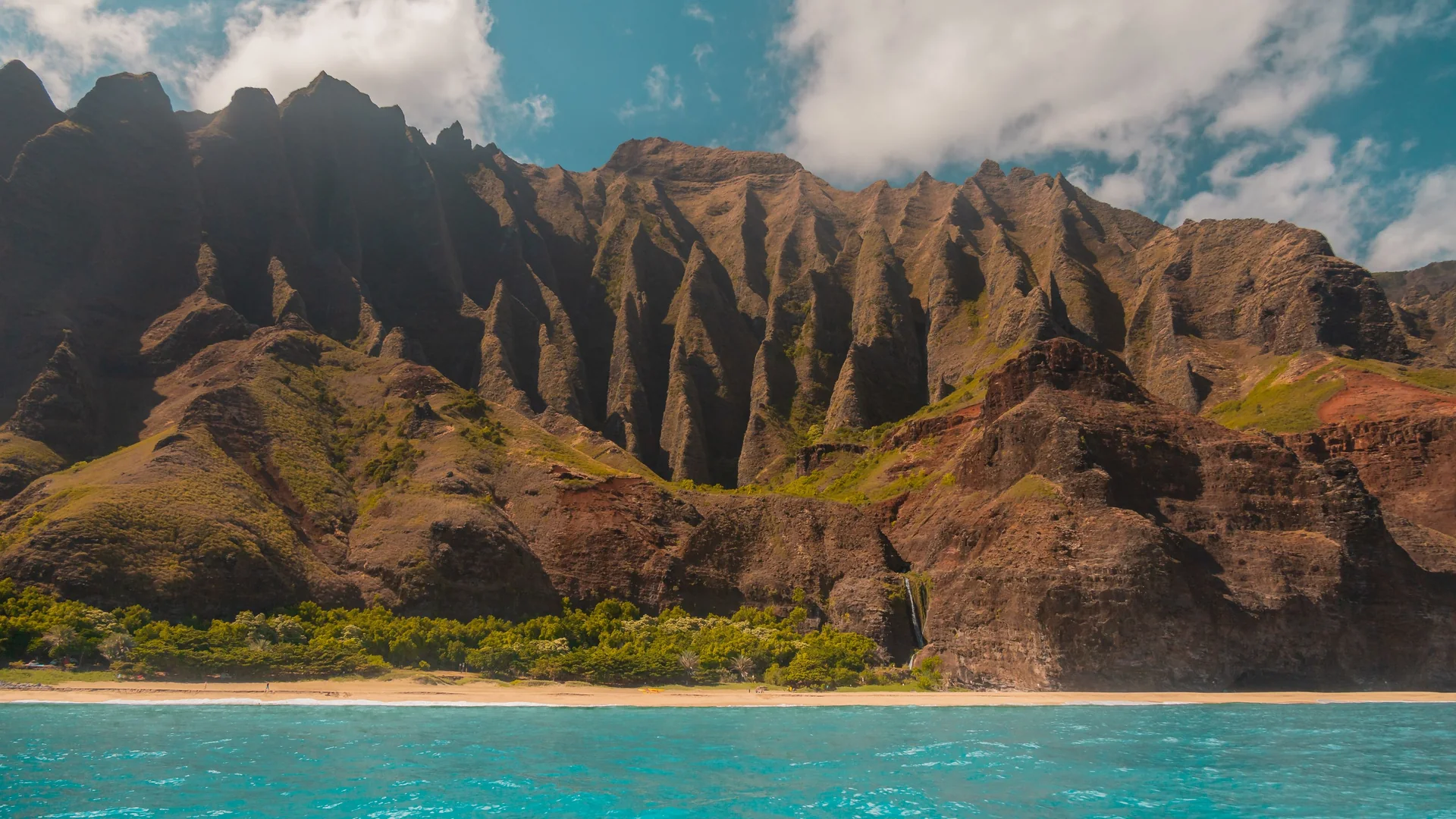 Fluted dark-red and green sea cliffs on Kauaʻi's Nāpali Coast rising behind a strip of sand and turquoise water, with a thin waterfall falling down the rock on the right