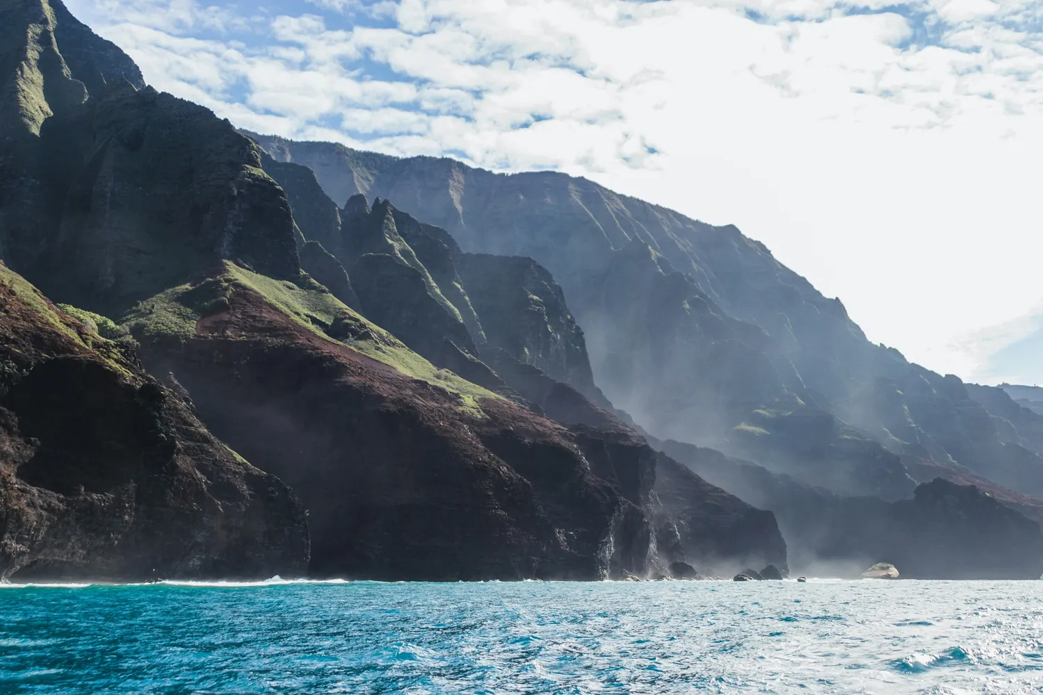 Dark Nāpali sea cliffs wrapped in sea mist, with waterfalls at their base, seen from the water