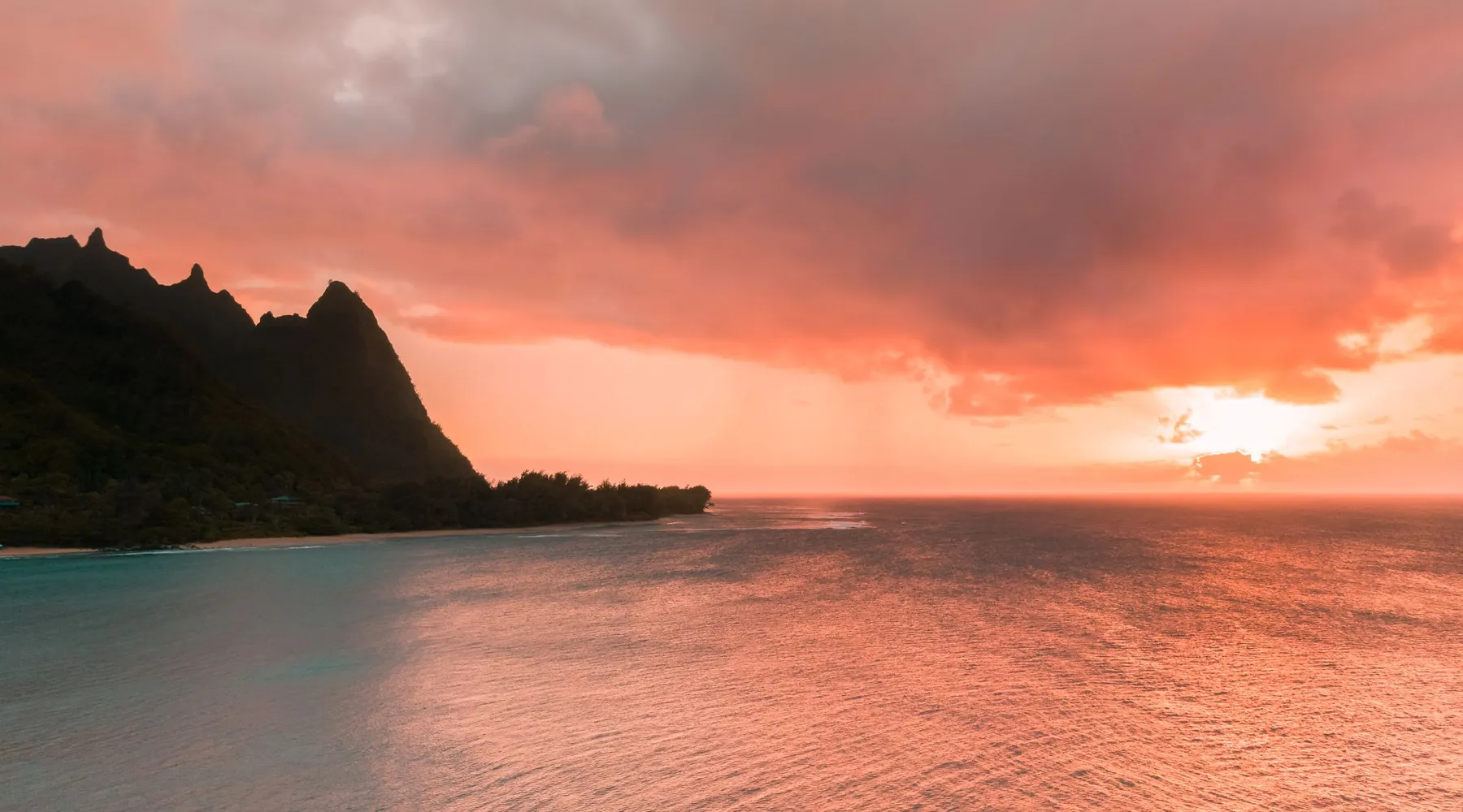 A coral-pink sky with the sun low on the horizon over calm water, and a dark ridge in silhouette on the left, on Kauaʻi's north shore
