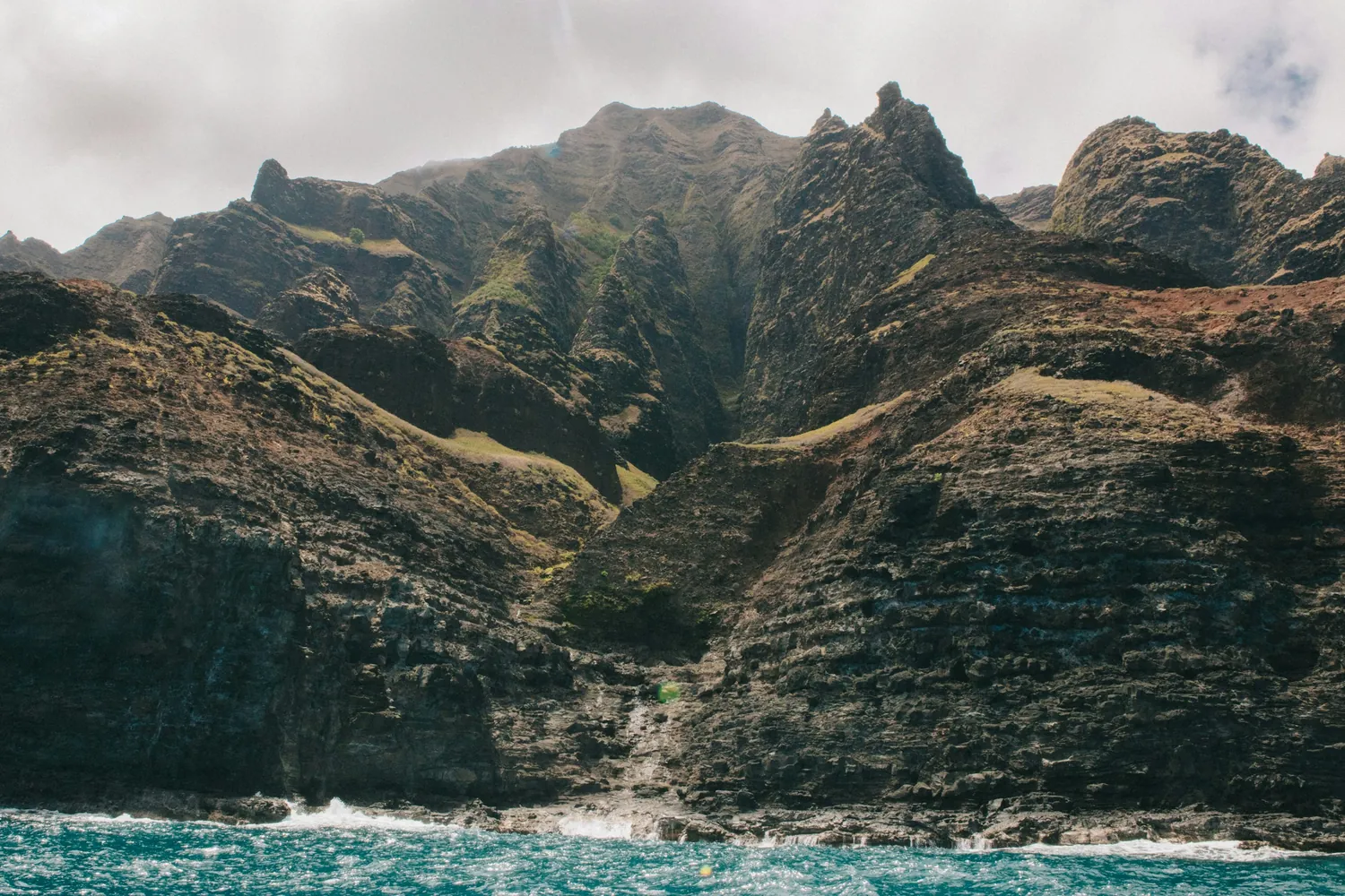 Layered Nāpali cliffs and a ridge crest rising above turquoise water, with a thin waterfall down the rock at the centre